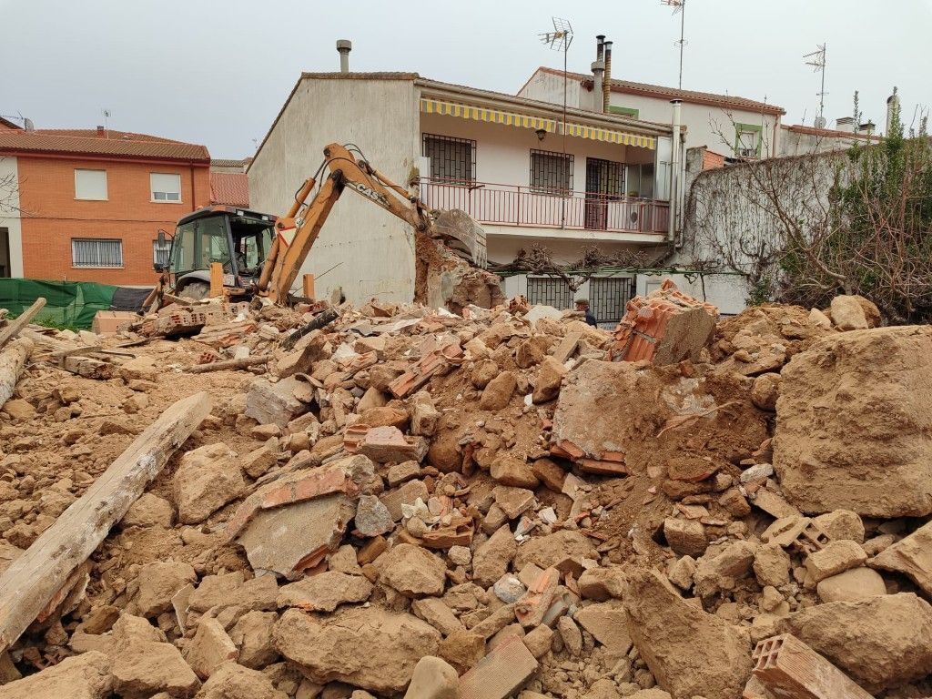 Obra de demolición con excavadora entre montones de escombros frente a un edificio parcialmente derribado.