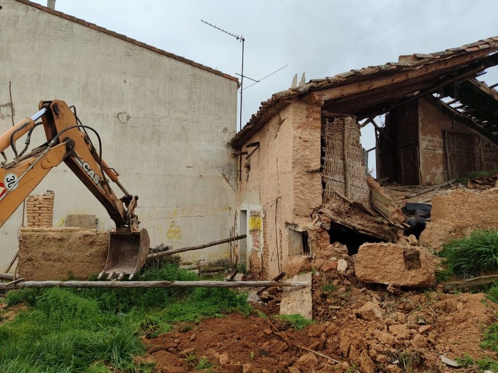Una excavadora demuele una casa de ladrillos parcialmente derrumbada junto a un muro liso.