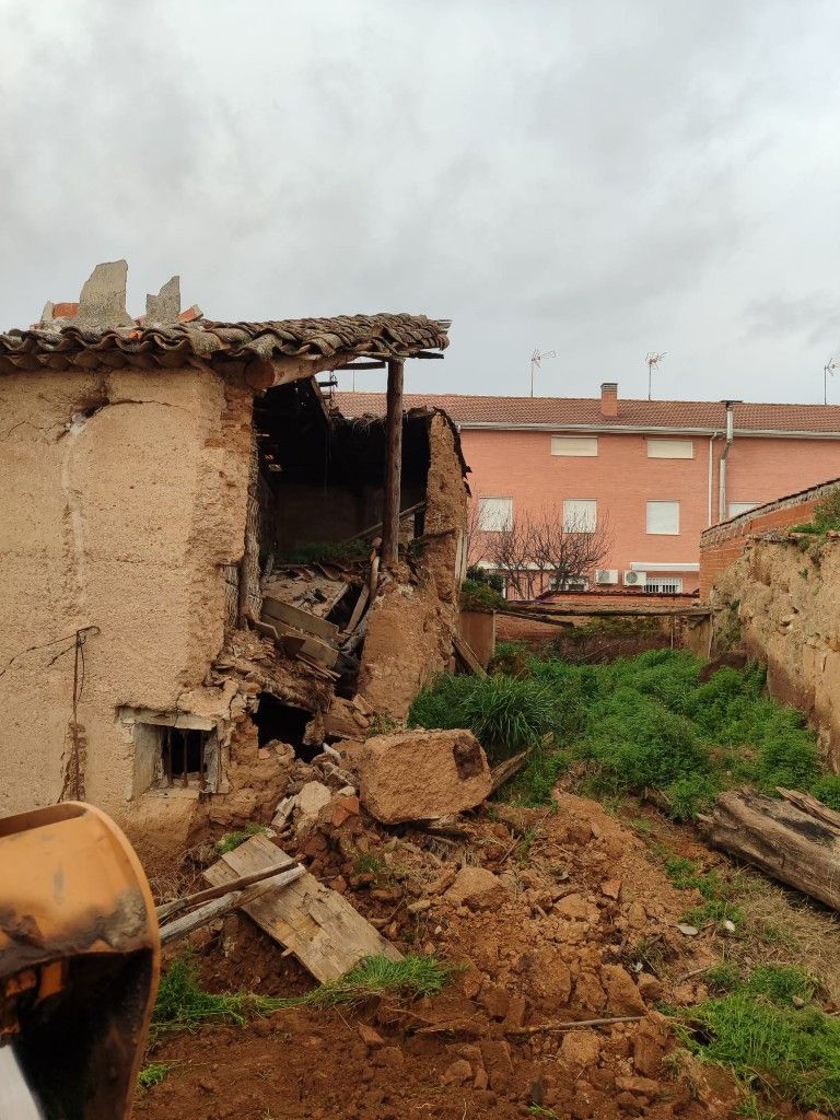 Edificio de adobe derrumbado con escombros en primer plano y una casa rosa al fondo bajo un cielo nublado.