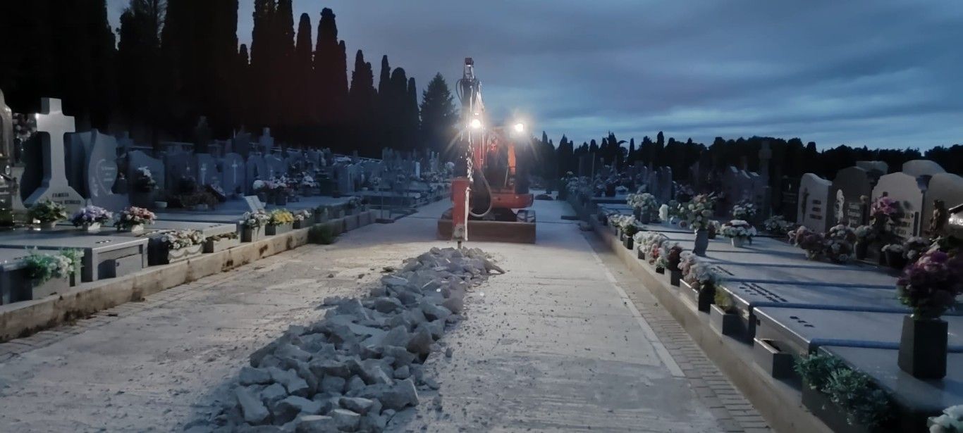 Sendero nevado del cementerio al atardecer, con hileras de lápidas y luces distantes bajo un cielo oscuro.