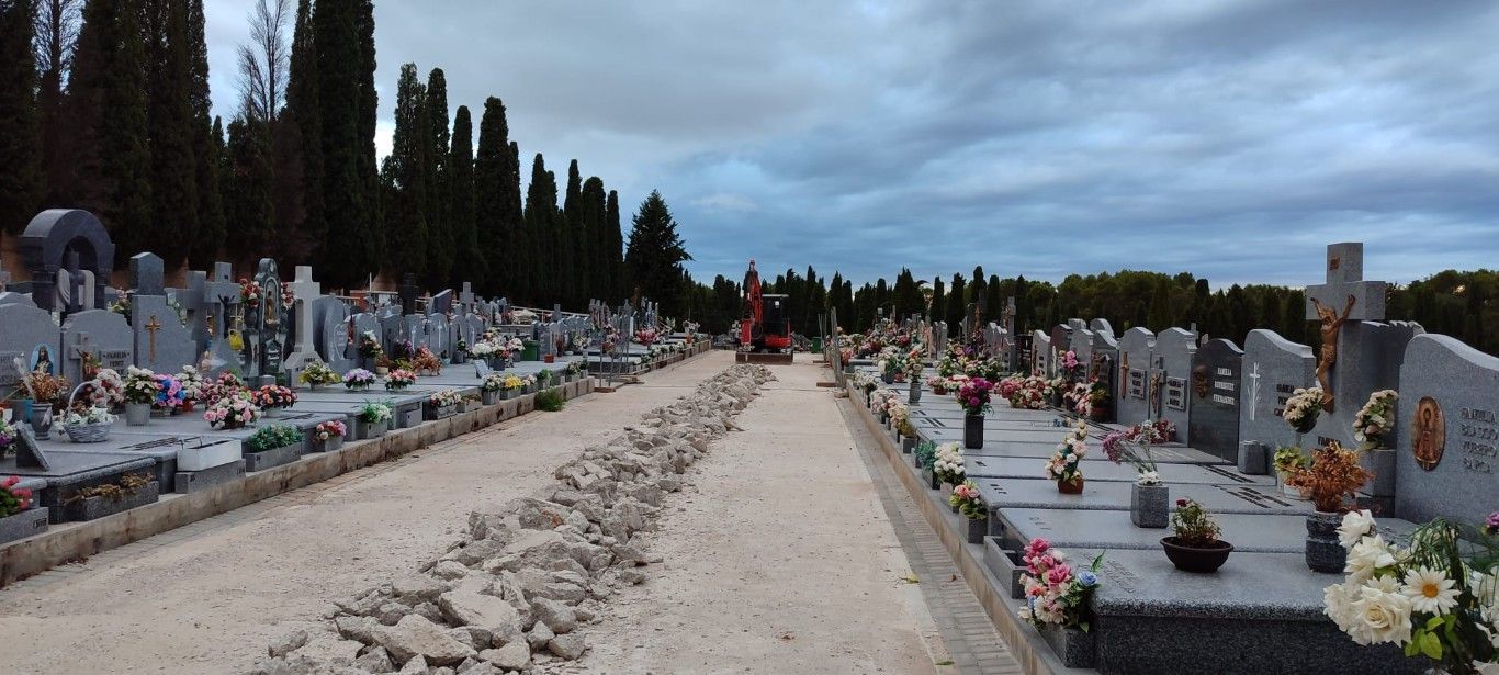 Amplio cementerio con hileras de tumbas y flores a lo largo de un sendero central de grava bajo un cielo nublado.