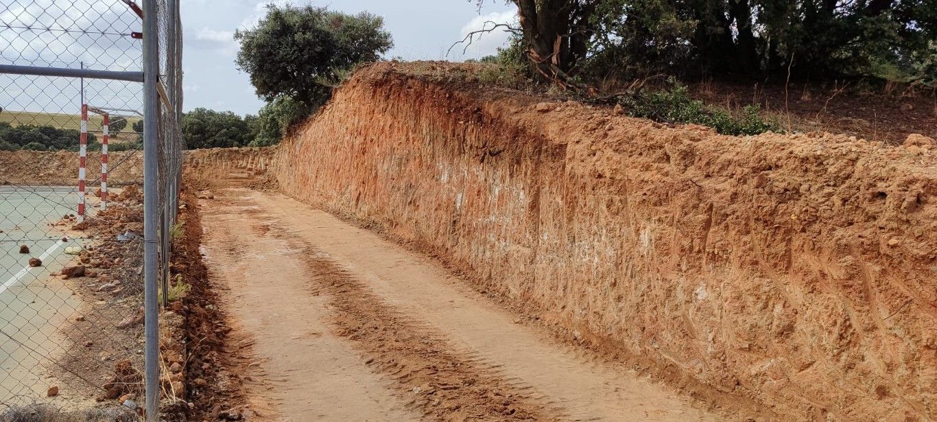 Sendero de tierra junto a un terraplén de color marrón anaranjado y una valla de tela metálica bajo un cielo despejado.