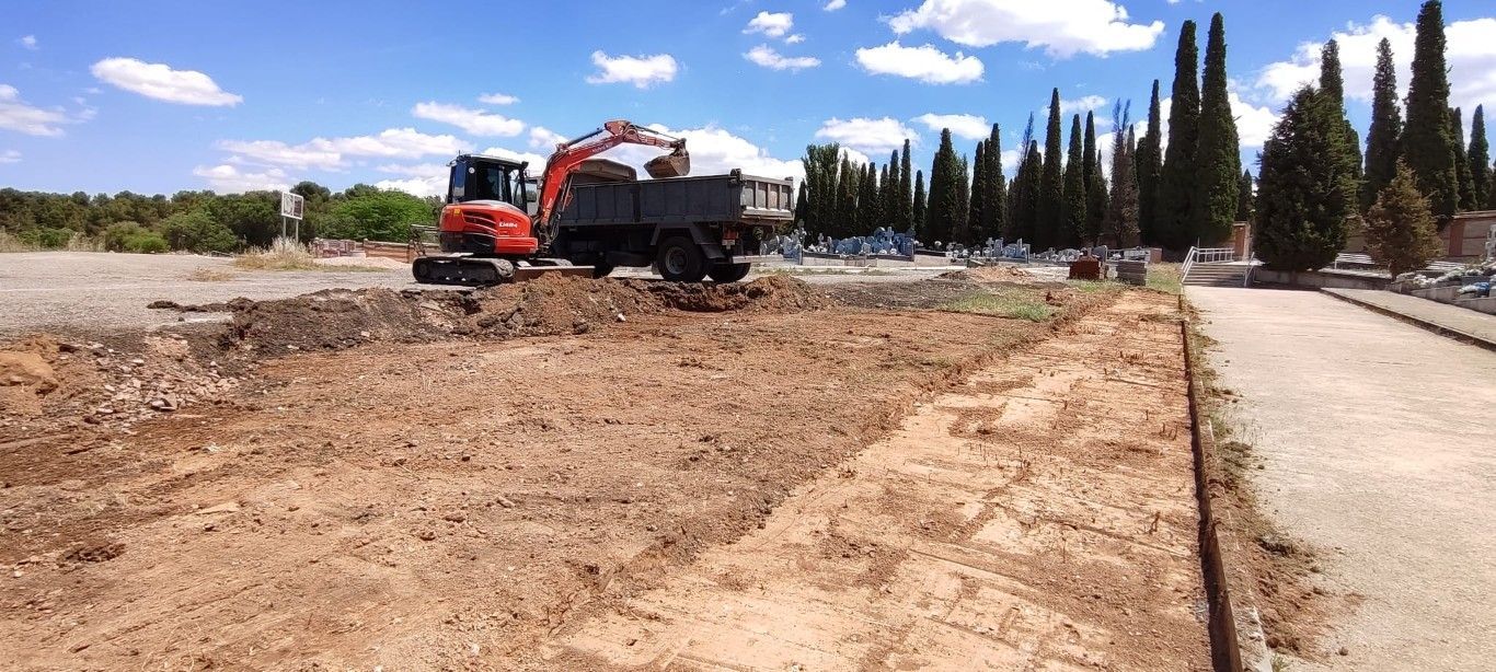 Excavadora roja y camión volquete en un terreno baldío junto a una carretera asfaltada con árboles altos bajo un cielo azul.