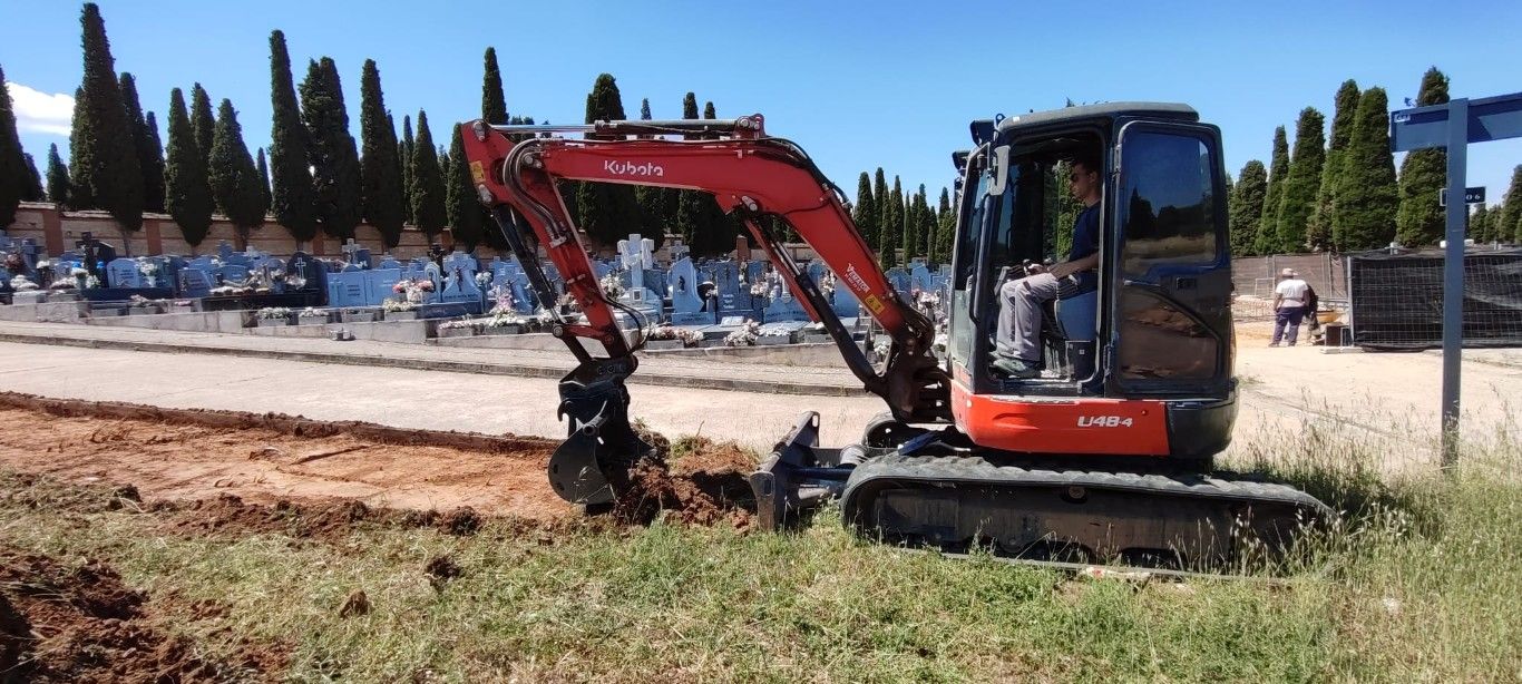 Excavadora roja excavando junto a una carretera, con árboles y una multitud al fondo.