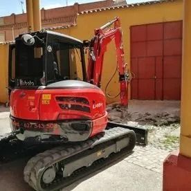 Una miniexcavadora roja aparcada en un patio junto a un muro amarillo y una puerta roja.