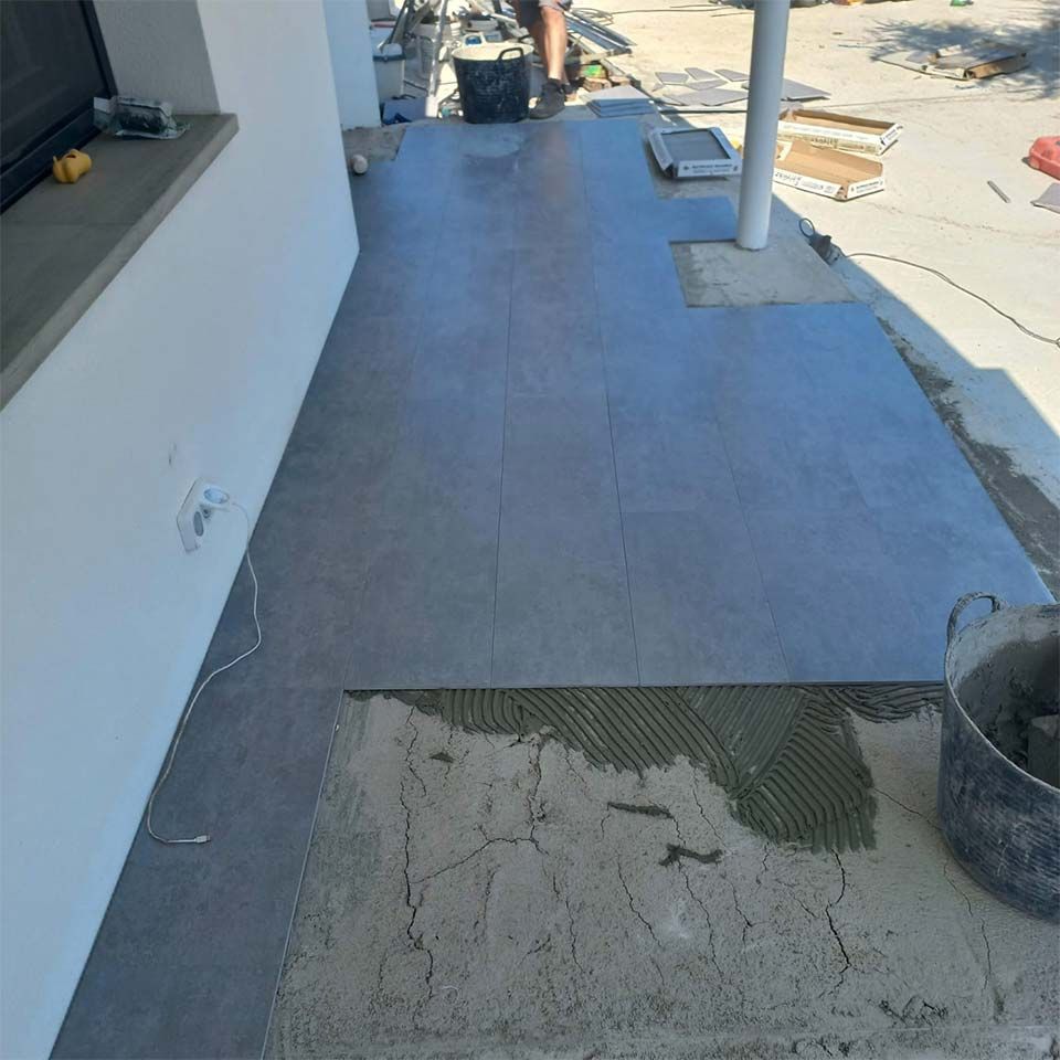 Alicatando un patio; un trabajador coloca baldosas grises. Cemento y herramientas visibles. Exterior, luz solar.