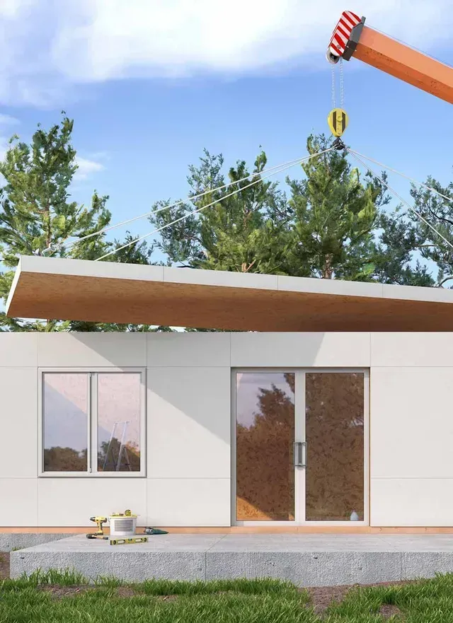 Grúa colocando un panel de techo en una casa modular prefabricada con revestimiento blanco, puerta de vidrio y una ventana.
