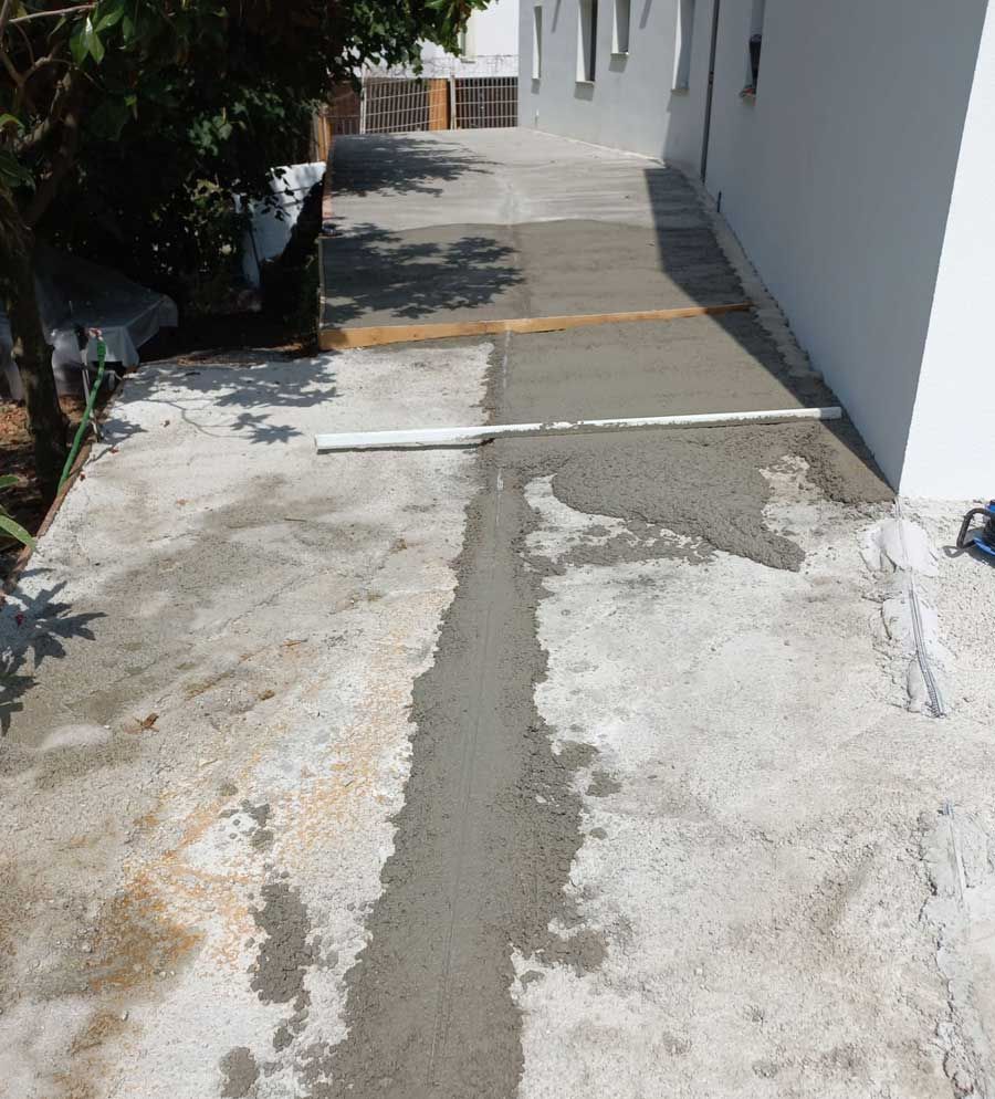 Camino de concreto en reparación; una franja de concreto nuevo contrasta con concreto más antiguo y teñido, con una barra niveladora y un edificio blanco.
