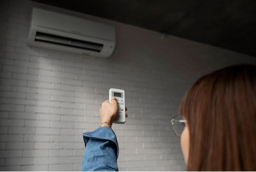 Mujer usando el control remoto del aire acondicionado en una habitación con pared de ladrillo blanco.