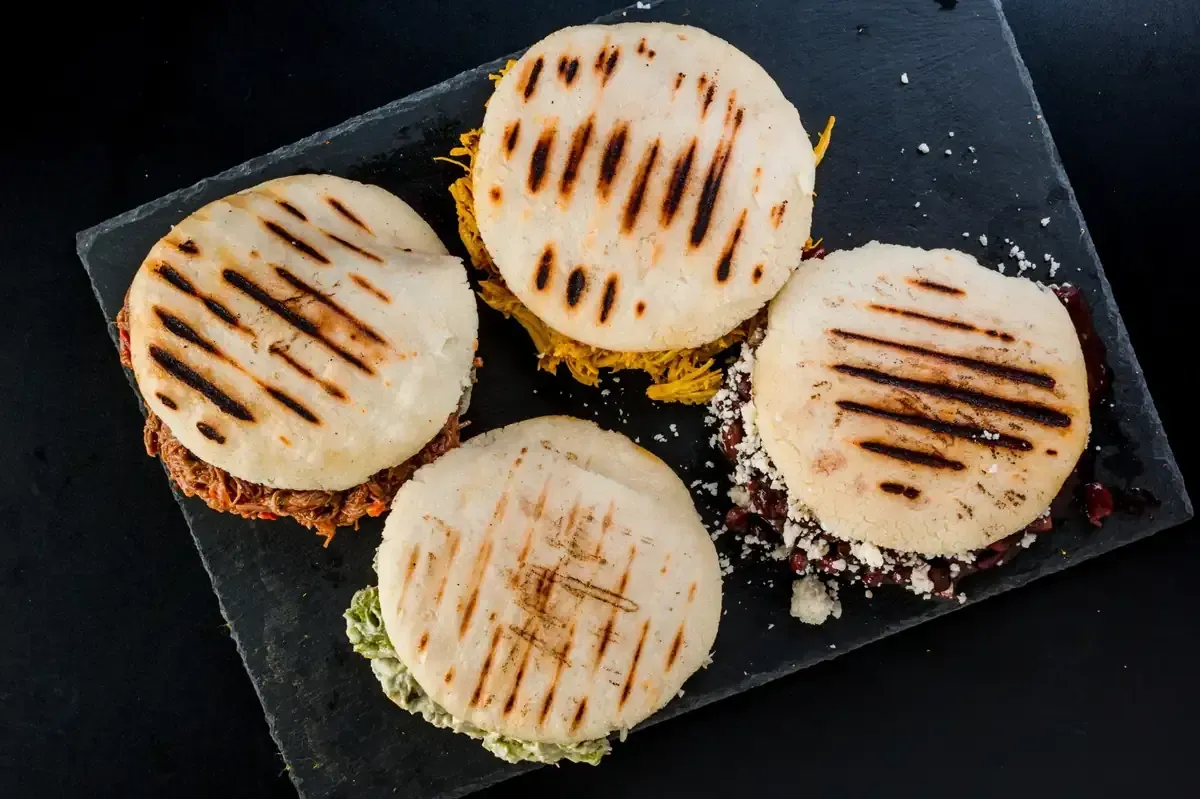 Cuatro arepas a la parrilla con diversos rellenos sobre una pizarra negra.