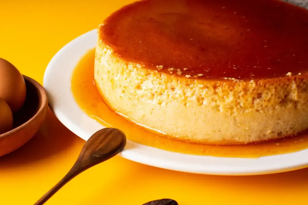 Flan en plato blanco con salsa de caramelo, cuchara de madera y huevos, sobre superficie amarilla.