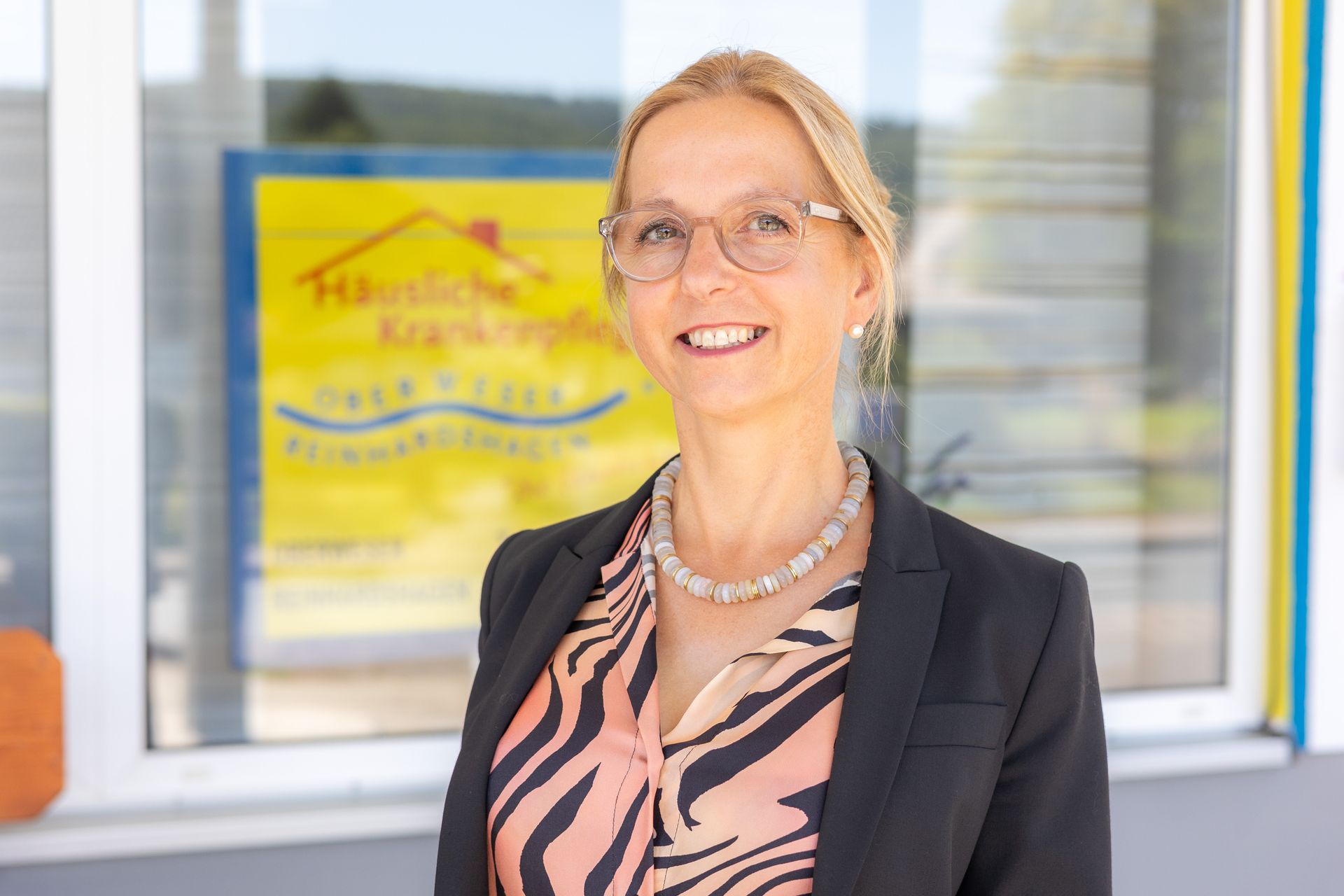 Eine Frau mit Brille und Blazer lächelt vor einem Fenster mit einem Logo.