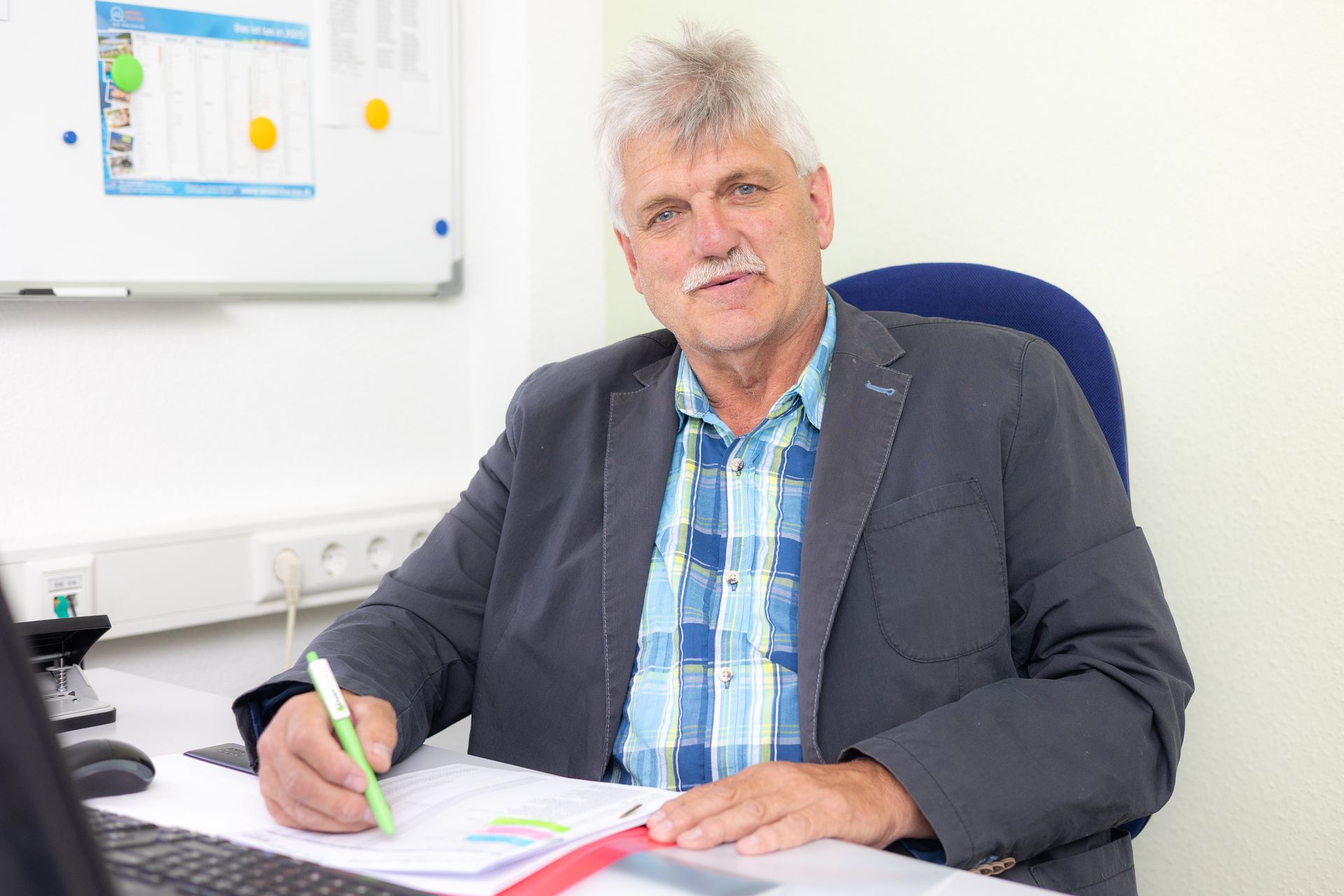 Mann im Blazer am Schreibtisch, schreibend. Im Hintergrund eine weiße Tafel.