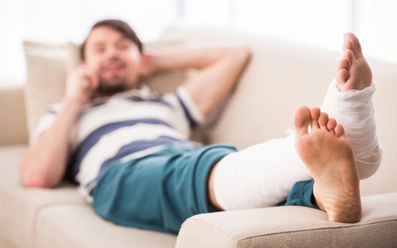 Un homme assis sur un canapé, la jambe et le pied plâtrés, parle au téléphone.