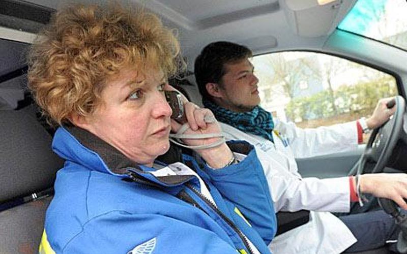 Une femme en veste bleue, au téléphone, à côté d'un médecin au volant ; à l'intérieur d'un véhicule.