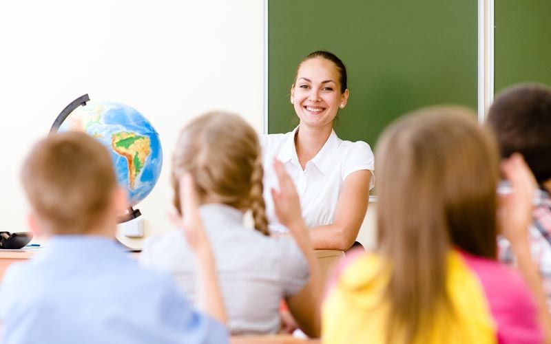 Une enseignante sourit à ses élèves dans une salle de classe. Plusieurs enfants lèvent la main ; on aperçoit un globe terrestre et un tableau noir.