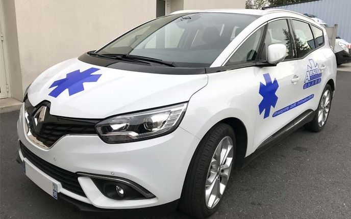 Ambulance Renault Scenic blanche avec des étoiles médicales bleues sur le capot et les côtés, garée devant un bâtiment.