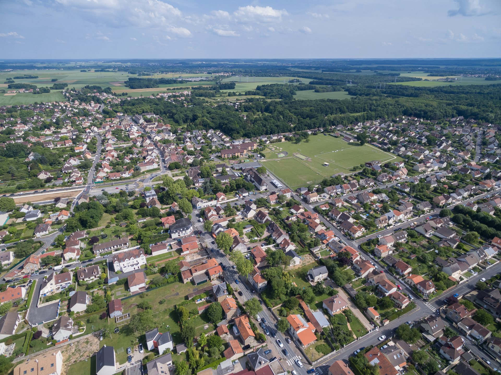 Vue aérienne d'une ville avec des maisons, des routes, des champs verdoyants et des arbres environnants sous un ciel bleu.