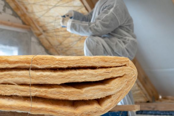 Des piles d'isolant au premier plan, un ouvrier en combinaison de protection installe de l'isolant dans les combles.