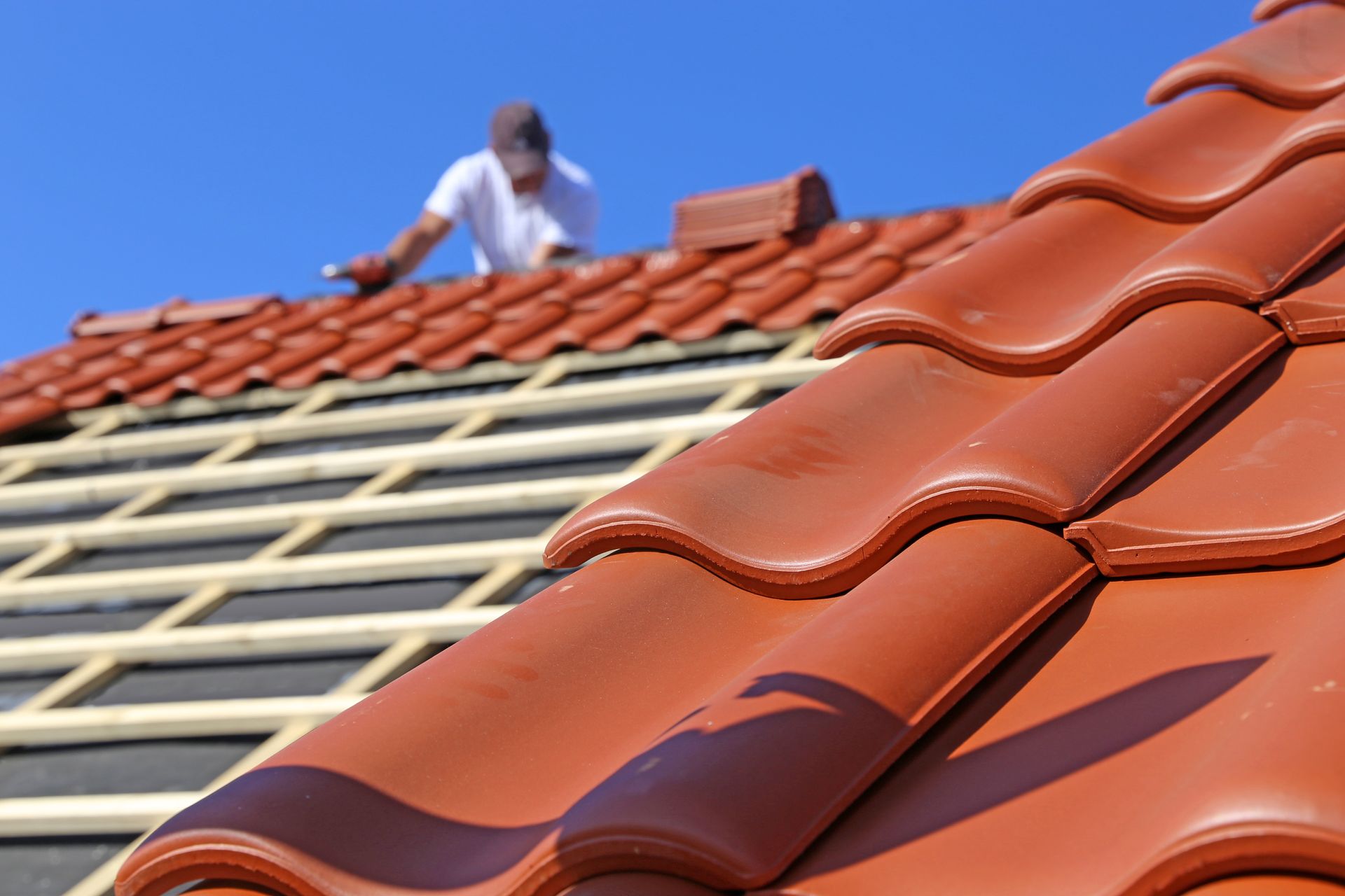 Un couvreur pose des tuiles de céramique brun rougeâtre sur une maison sous un ciel bleu dégagé.