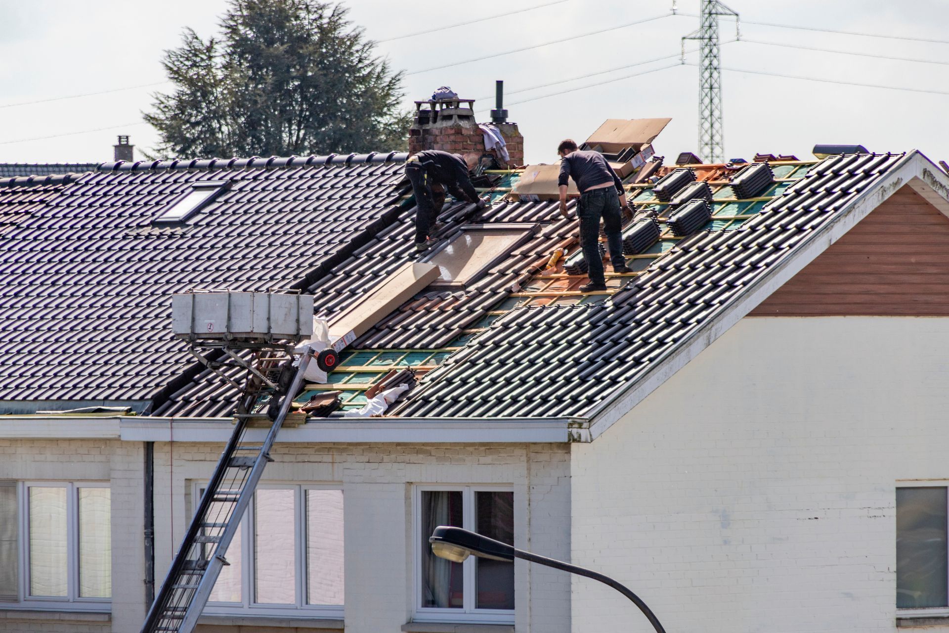 Des couvreurs réparent un toit de tuiles sur un bâtiment résidentiel, à l'aide d'une échelle et travaillant sur le toit sous un ciel dégagé.