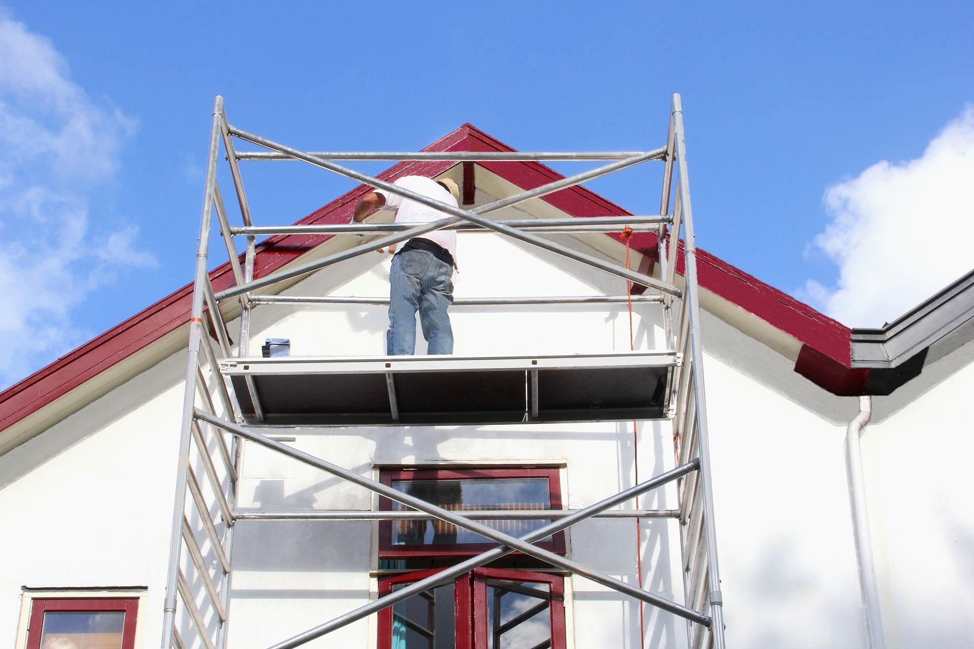 Une personne sur un échafaudage peint le toit d'un bâtiment blanc à bordures rouges sur fond de ciel bleu.