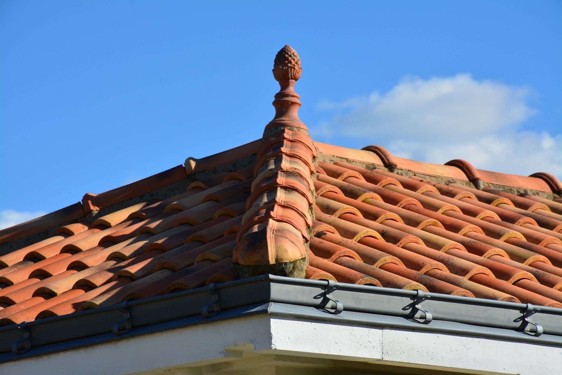Toit de tuiles rouges orné d'une arête et d'un fleuron décoratifs, se détachant sur un ciel bleu.