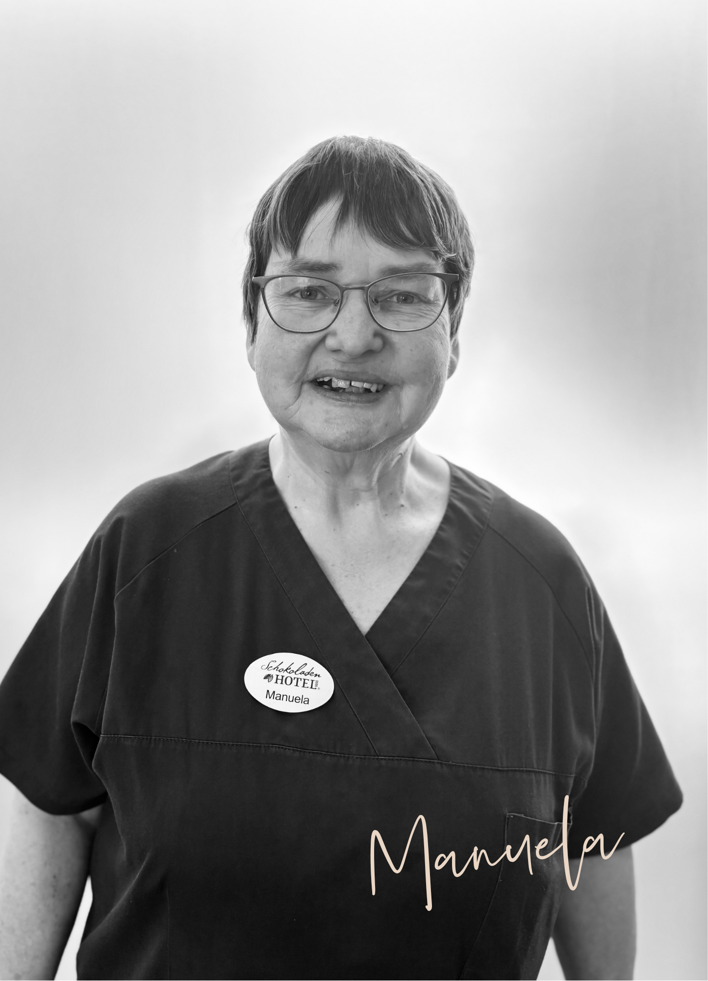 Eine Frau lächelt; sie trägt eine Brille und ein Namensschild. Ihre Unterschrift lautet „Manuela“.