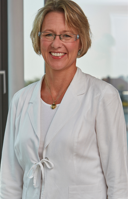 Eine Frau mit Brille und weißer Jacke lächelt in die Kamera