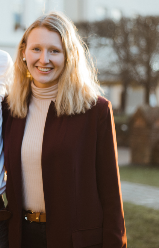 Eine Frau in einer kastanienbraunen Jacke lächelt in die Kamera
