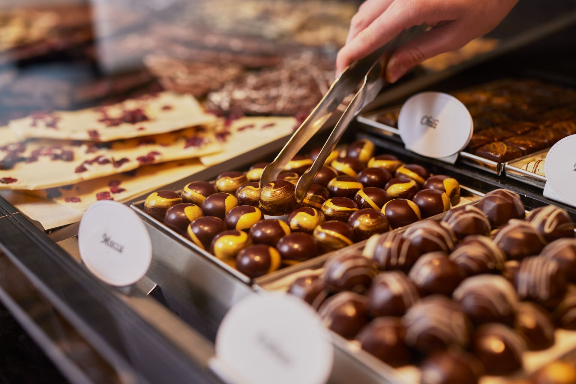 Eine Person nimmt mit einer Zange eine Praline aus einer Auslage