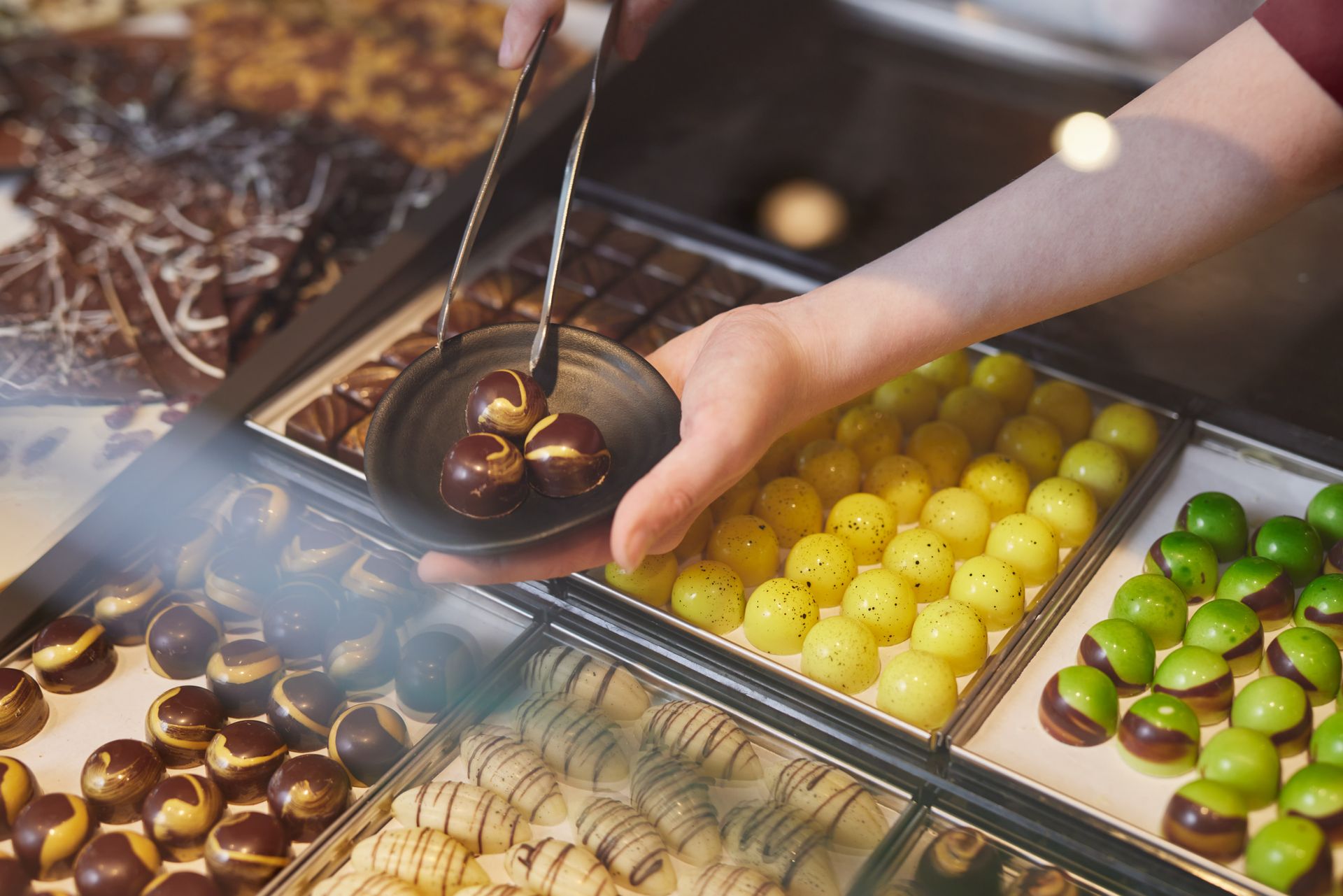 Eine Person nimmt sich mit einer Zange Pralinen aus einer Auslage und legt sie auf einen kleinen Teller.