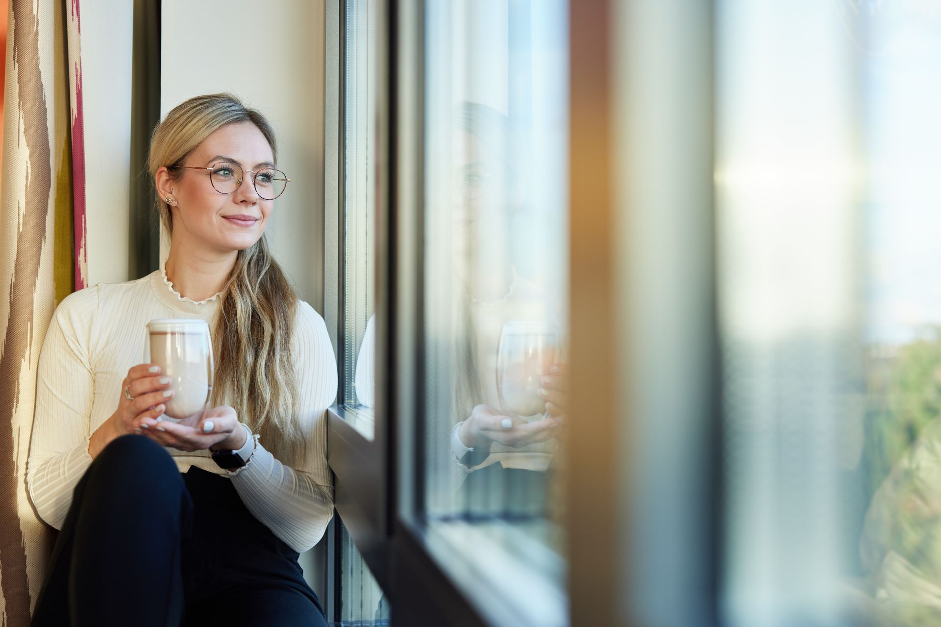 Eine Frau sitzt auf einem Fensterbrett, hält eine Tasse Kaffee und schaut aus dem Fenster.