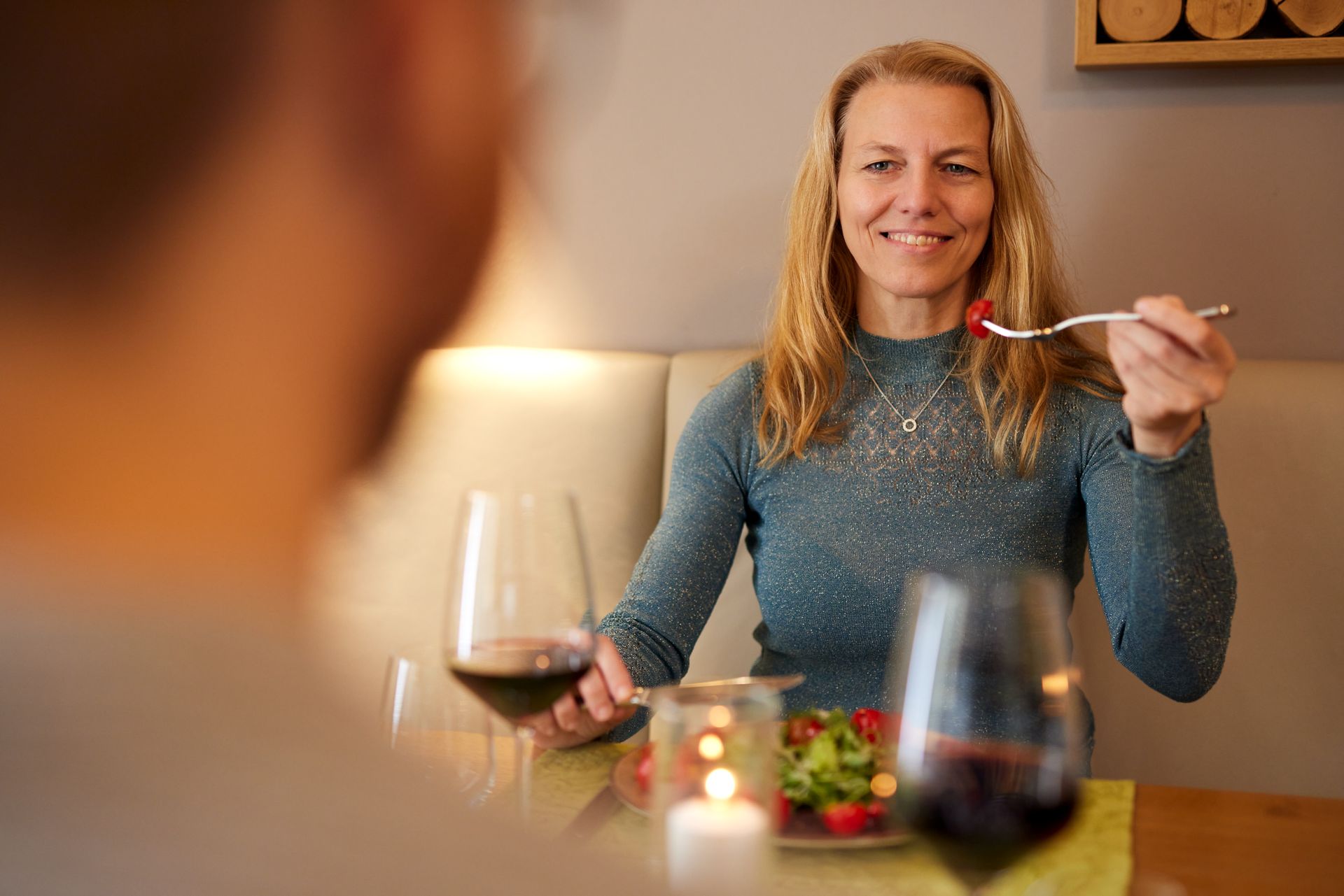 Frau isst Salat, lächelt der Person gegenüber am Tisch zu, hält eine Gabel, Weingläser und eine Kerze in der Hand.