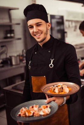 Ein Koch hält in einer Küche zwei Teller mit Essen.