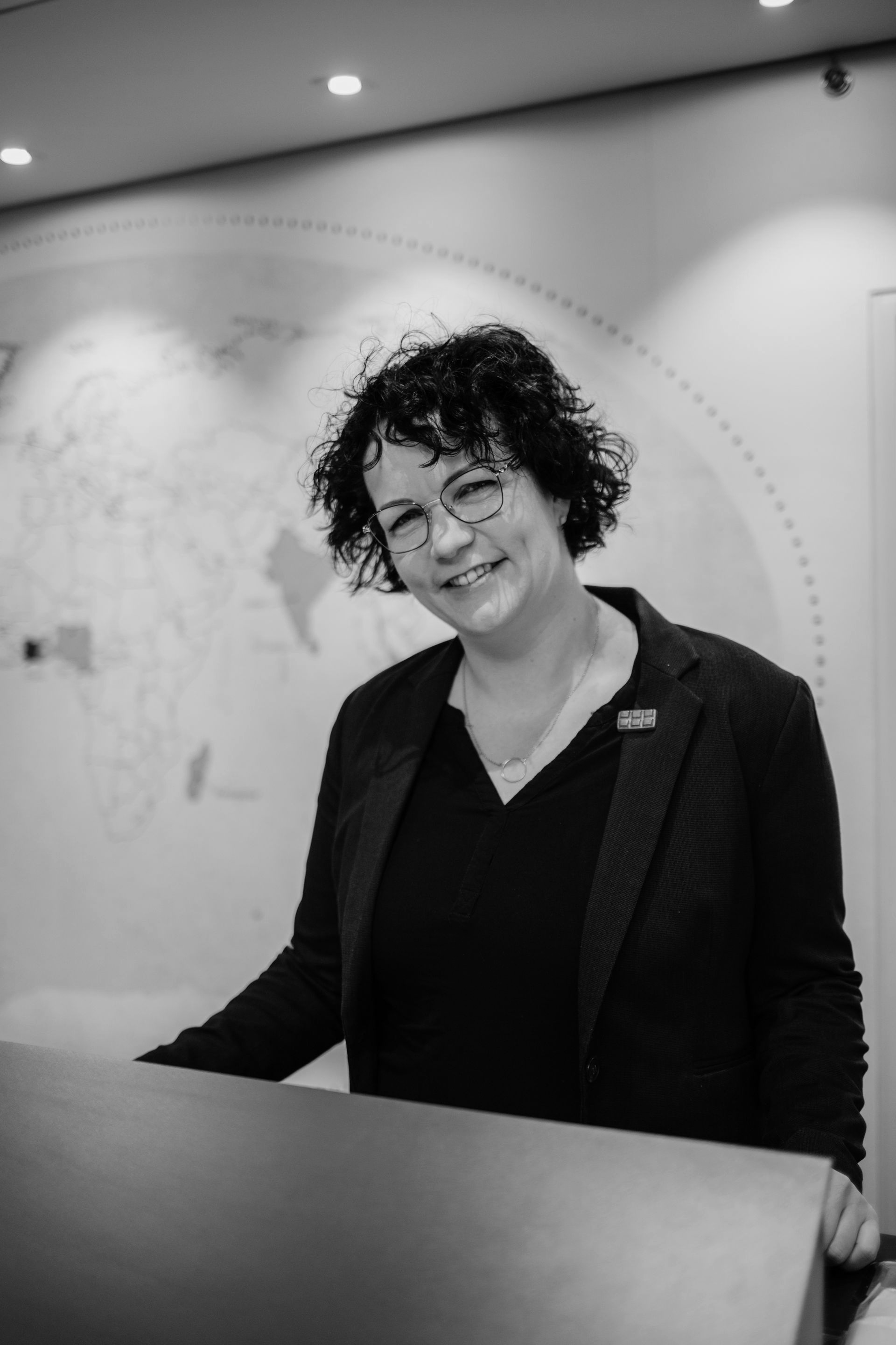 Eine Frau in einer schwarzen Jacke und mit Brille steht hinter einem Tresen.