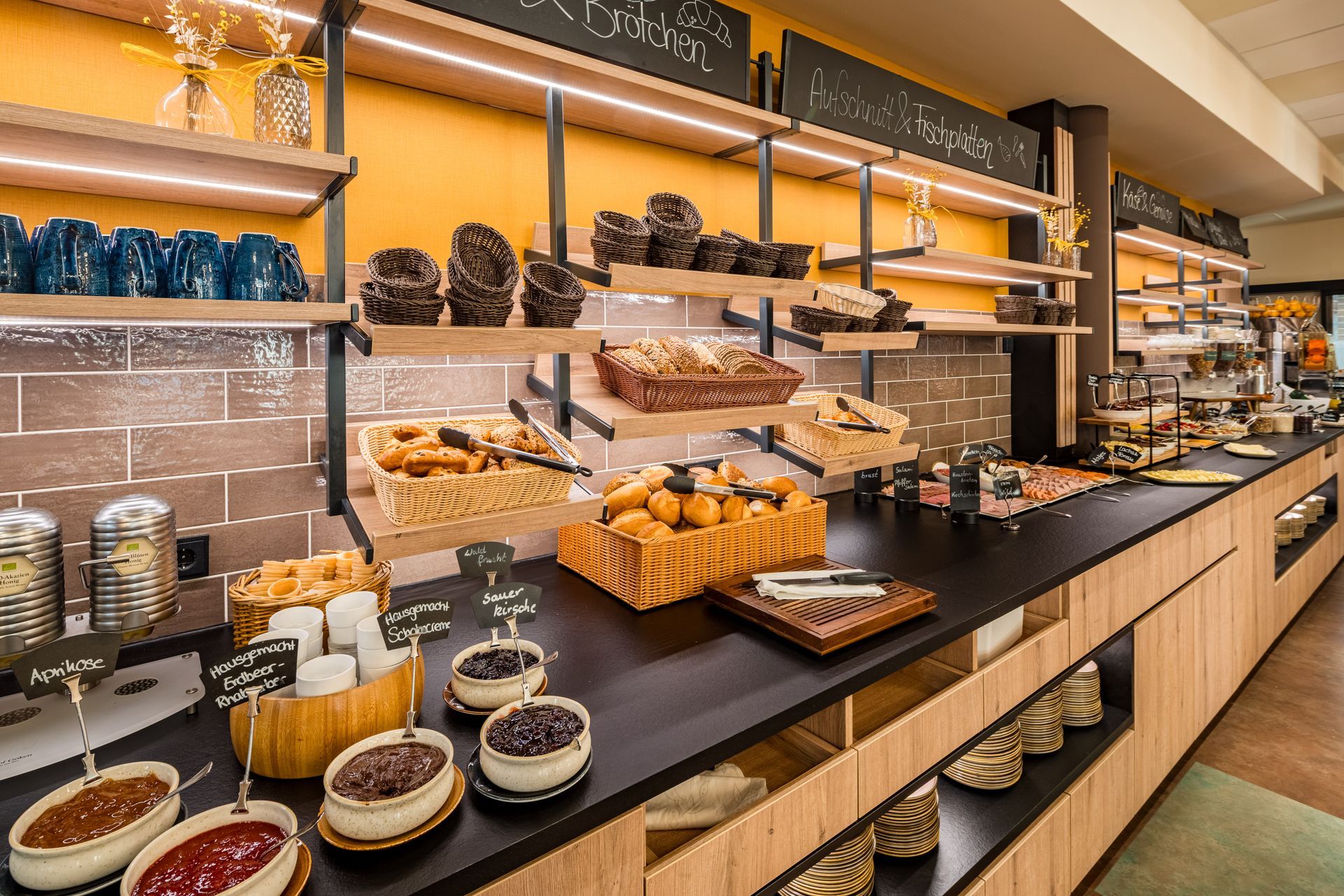 Buffet mit Gebäck, Marmeladen und Brot, präsentiert auf Holzregalen und -theken, in Gelb- und Brauntönen gehalten.