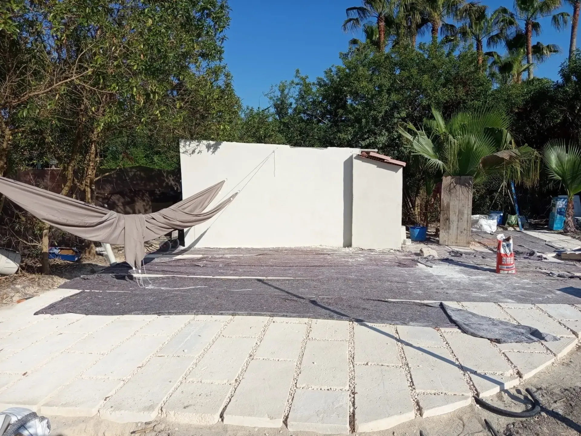Obreros construyendo una acera de hormigón junto a una casa, con árboles y materiales de construcción en las cercanías.