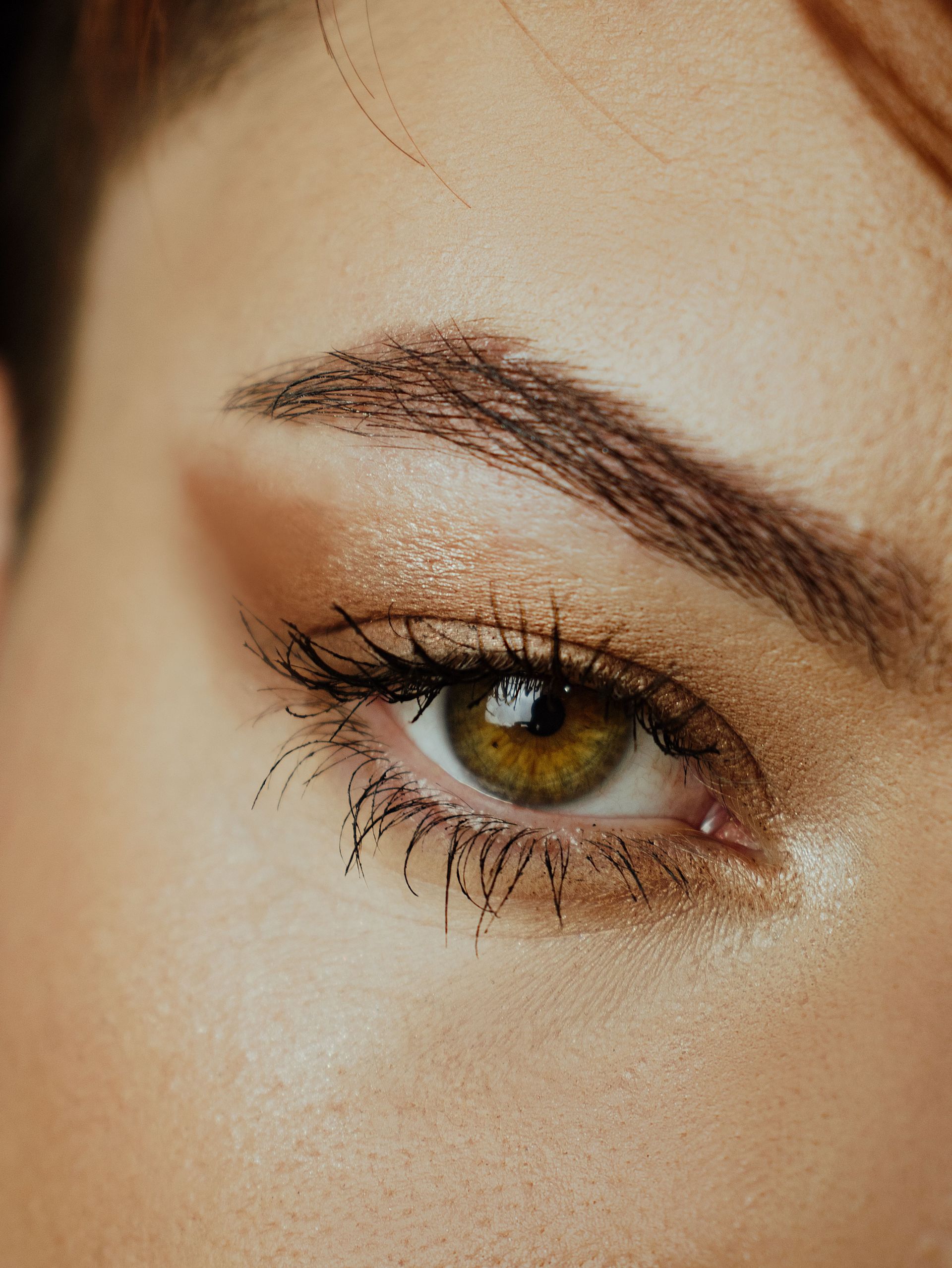 Une main tenant un pinceau de maquillage au-dessus d'un petit bol de crème beige dans une pièce lumineuse et tamisée