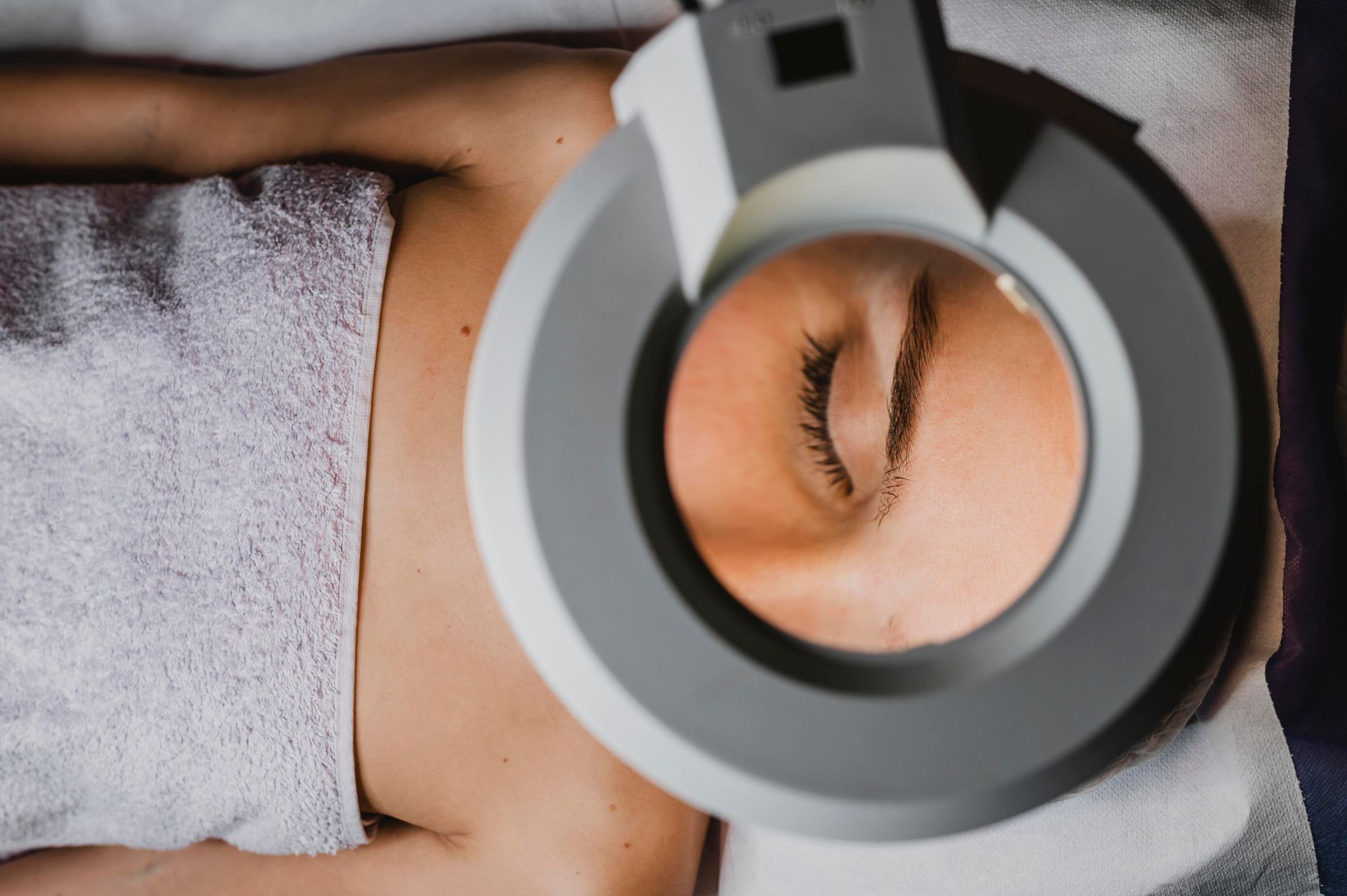 Gros plan d'une personne allongée, les yeux fermés, sous une lampe loupe dans un spa ou un salon de beauté.