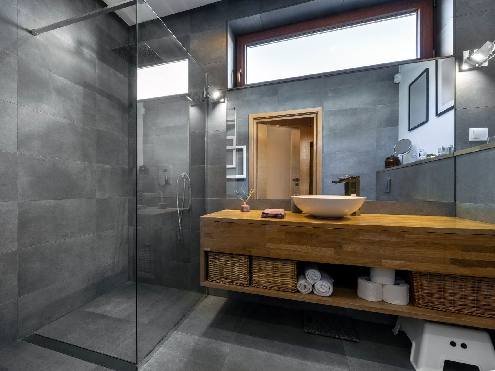 Salle de bains moderne avec murs carrelés gris, meuble-lavabo en bois et douche en verre.