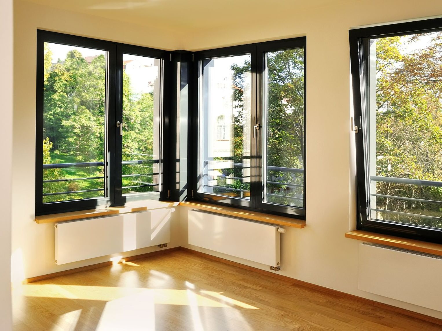 Chambre d'angle avec de grandes fenêtres à cadre noir donnant sur les arbres ; radiateurs blancs, parquet.