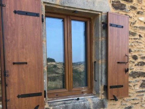 Fenêtre brune à volets sur un bâtiment en pierre. Les volets sont fermés et maintenus par des charnières et des ferrures noires.
