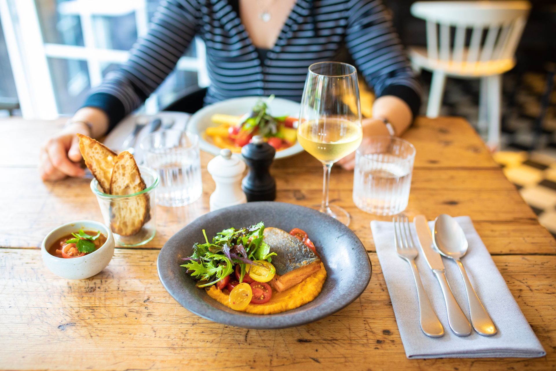 Eine Frau sitzt an einem Tisch mit Tellern voller Essen und einem Glas Wein.