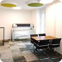Kleiner Besprechungsraum mit Drucker, Tisch und schwarzen Stühlen auf gemustertem Teppichboden.