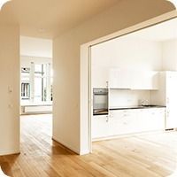 Helles, leeres Hausinterieur mit Holzböden, das sich zu einer weissen Küche und einem sonnendurchfluteten Raum öffnet.