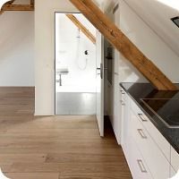 Moderne Küche mit weissen Schränken, Holzboden, schrägem Deckenbalken und offener Tür zu einem weissen Badezimmer.