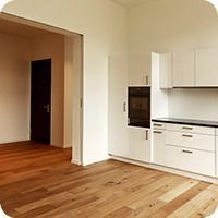 Leere, moderne Küche mit weissen Schränken und Holzboden, die sich zu einem angrenzenden Raum öffnet
