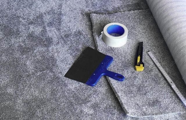 Werkzeuge zur Teppichreparatur auf grauem Teppich: blauer Spachtel, Klebebandrolle, Universalmesser und Lineal.