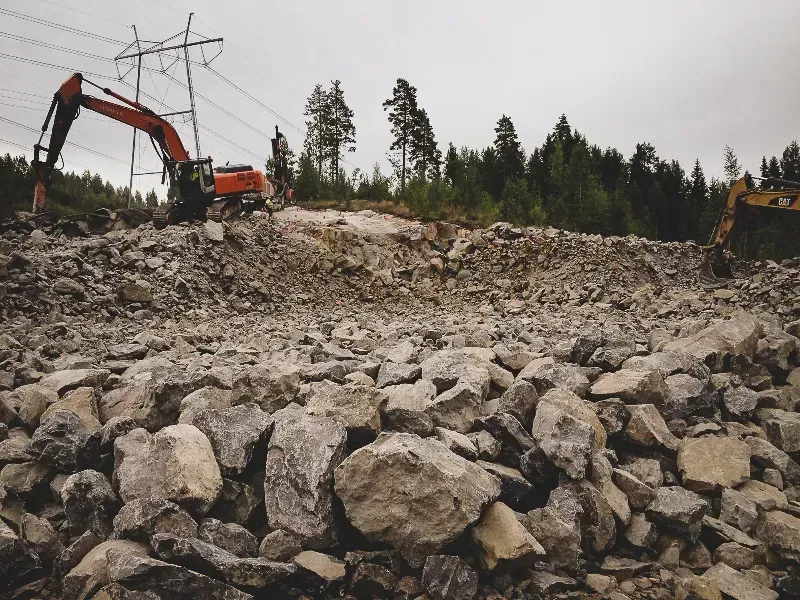 Kaivinkoneet työskentelevät kivisellä rakennustyömaalla voimalinjojen alla, taustalla metsä